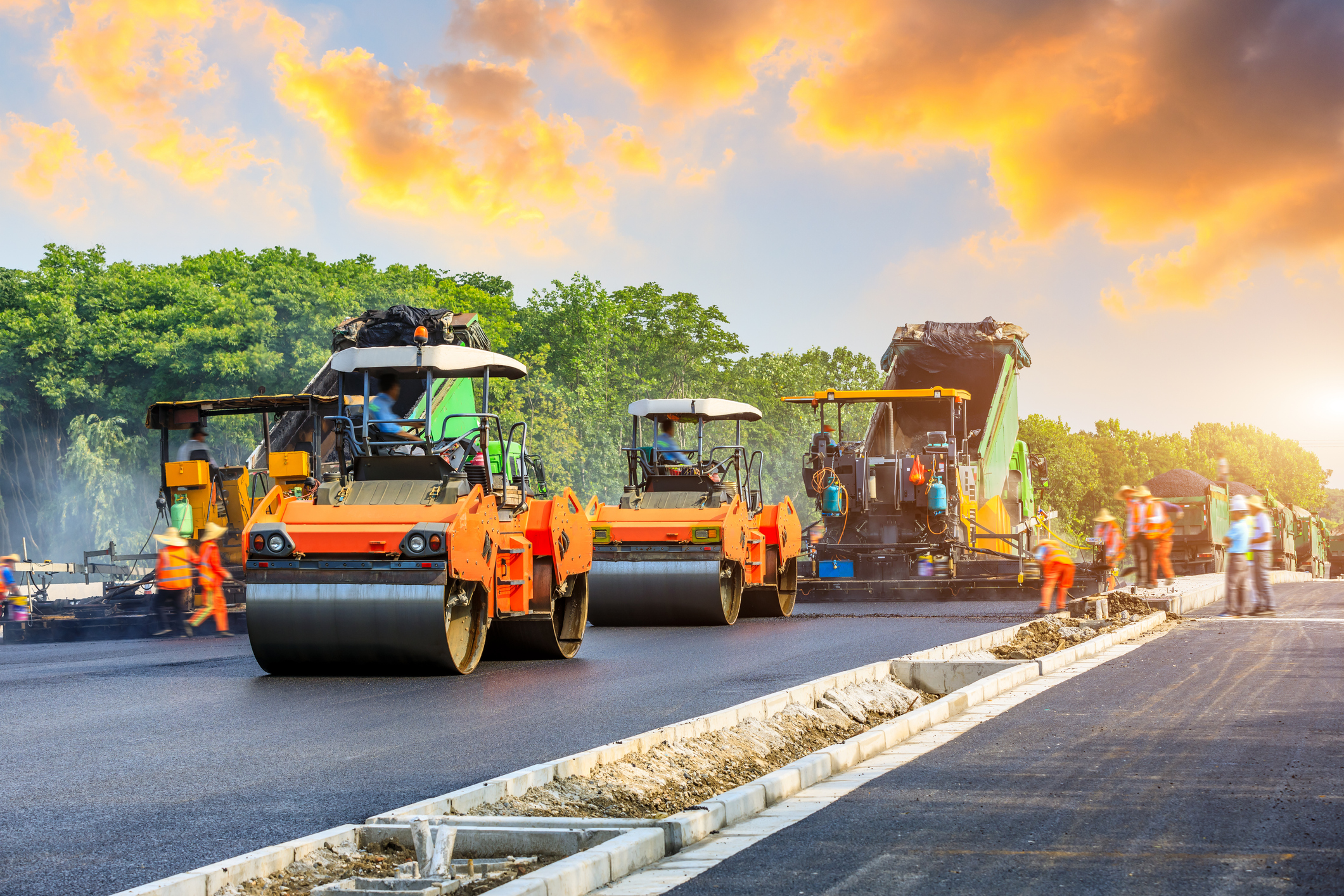 Road rollers and paving equipment resurfacing a road at sunset.